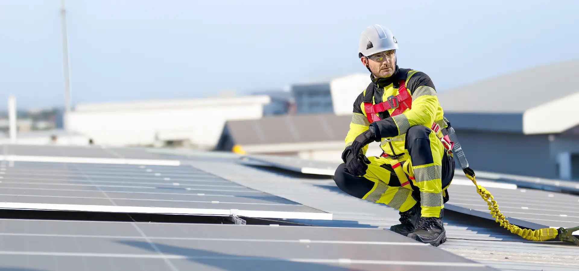 A worker in a high-visibility yellow and black safety suit with reflective stripes, wearing a white hard hat and gloves, crouches on a rooftop while holding a bright yellow fall arrest lanyard.1621243260e1af0c20-0